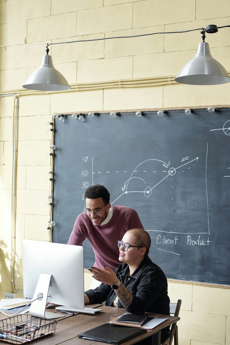 A man of colour standing next to a woman with a shaved head working at a computer and smiling.